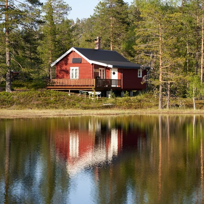 Charming red cabin reflected in a serene lake in Krøderen, Norway, amidst lush greenery.